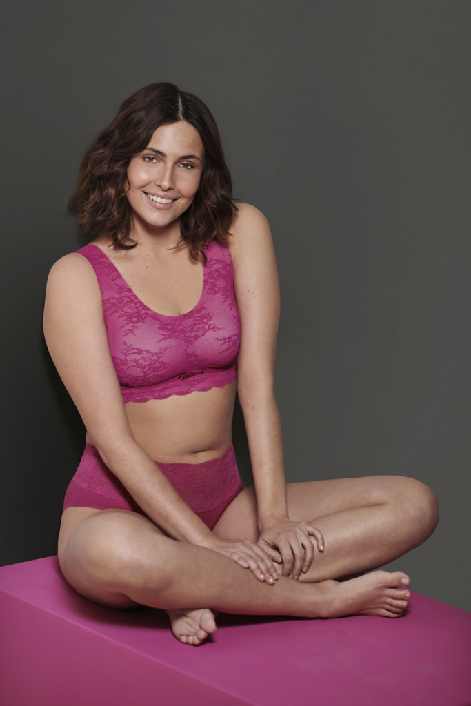 Dr. Anita Dr He sitzt auf einem pinken Podest und trägt ein pinkes Spitzen-Bralette mit passendem Slip